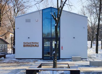 Auf dem Foto ist das Ausstellungsgebäude für die Werke Gerhard Richters von außen zu sehen. Das einfache Gebäude ähnelt einer Baracke und ist weiß gestrichten. Der Eingang ist zentral in der Mitte. An dieser Stelle ist bis zum Dach eine vertikale Fensterfront eingezogen.